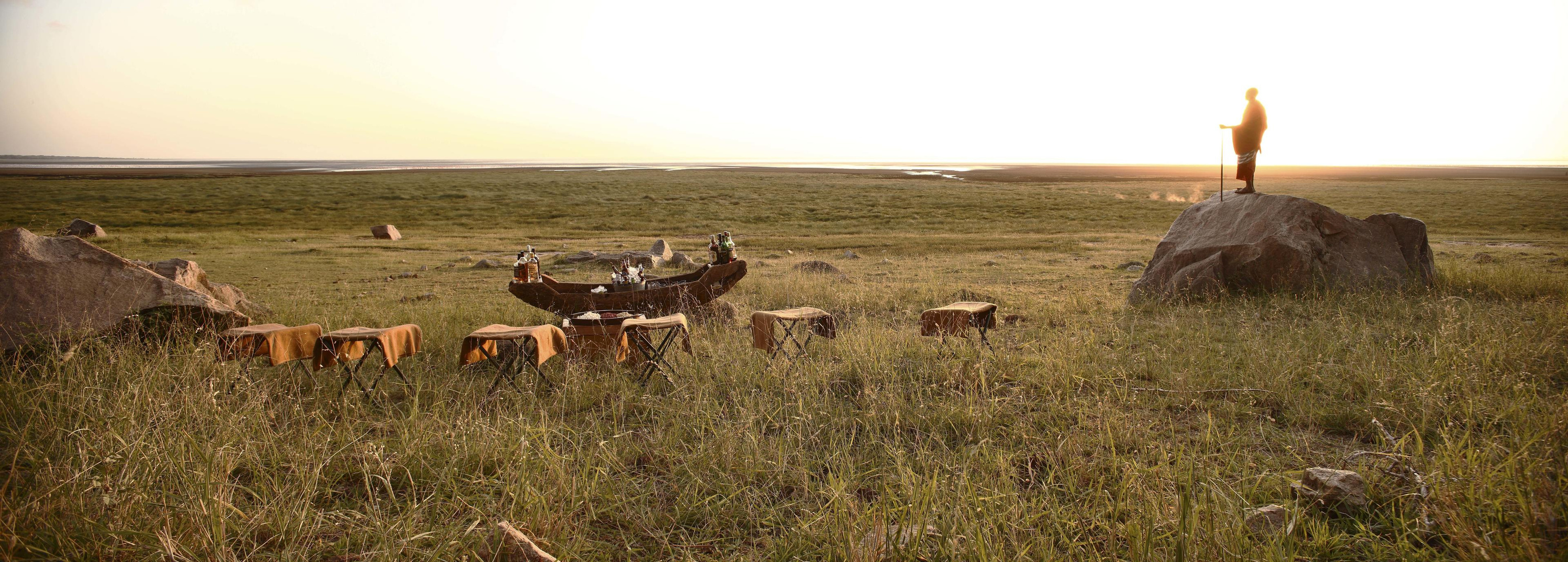 A sundowner set up on the East African plains at golden hour, a guide silhouetted on a rock.