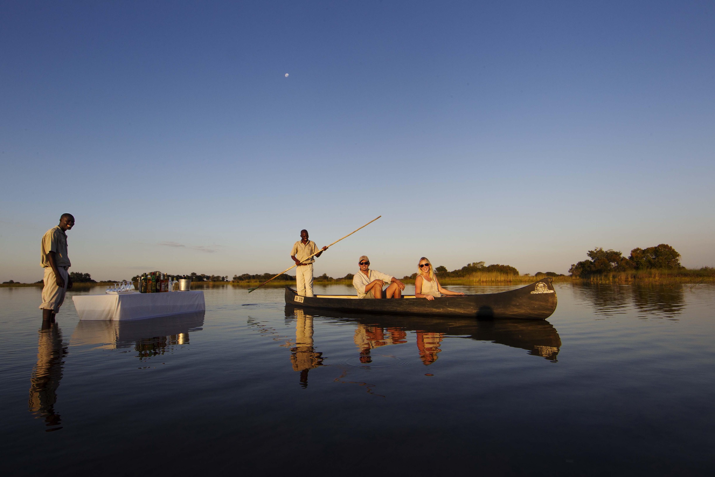 A sundowner table set on the still water of the Okavango Delta, mokoros at dusk.