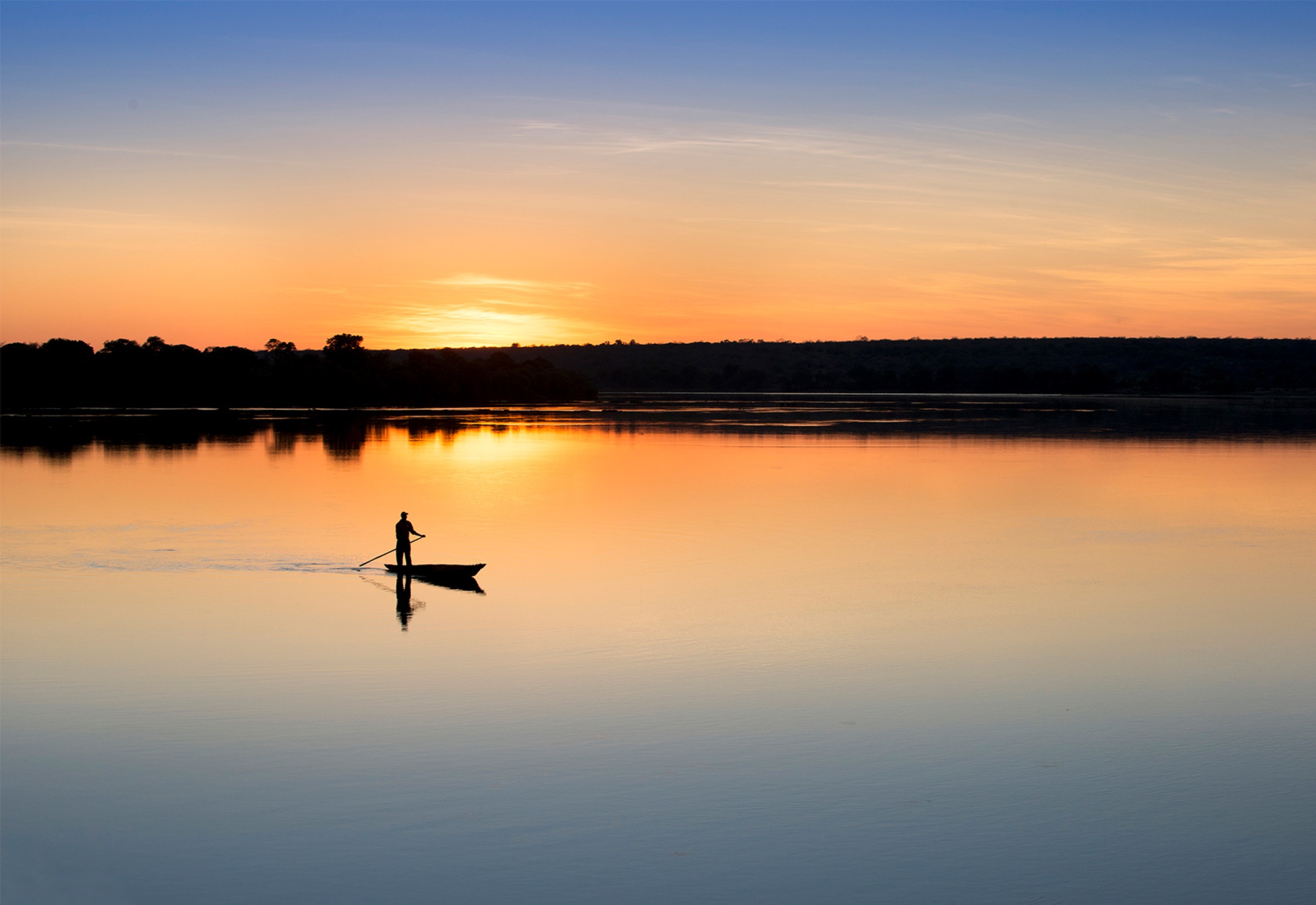 A lone mokoro paddler on a Zambezi-still river at sunset.